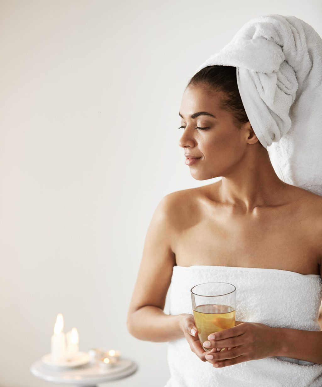 Beautiful african girl with towel on head holding glass smiling looking in side resting in spa resort. Copy space.