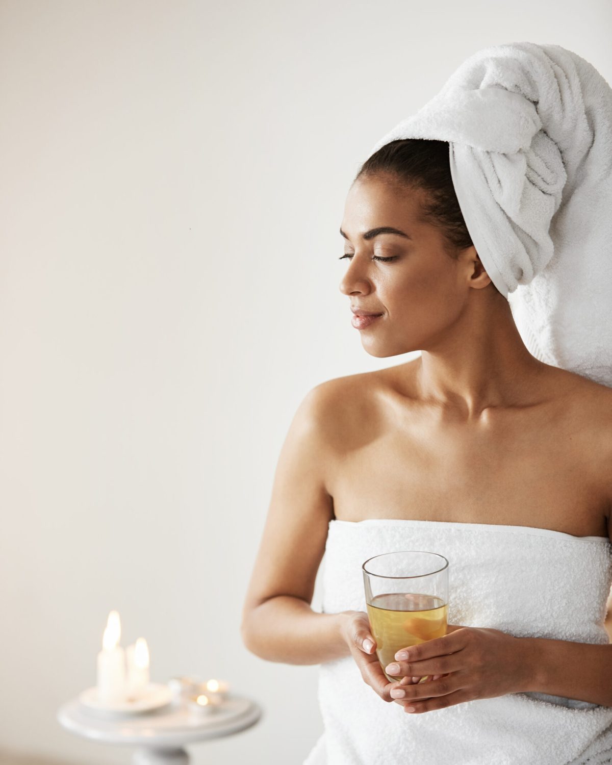 Beautiful african girl with towel on head holding glass smiling looking in side resting in spa resort. Copy space.